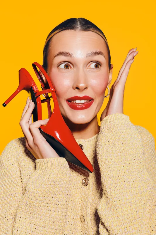 Young woman with red high heel shoe near face looks surprised and happy. Bright yellow background and cozy beige sweater create warm and playful mood. Concept of fashion and style.