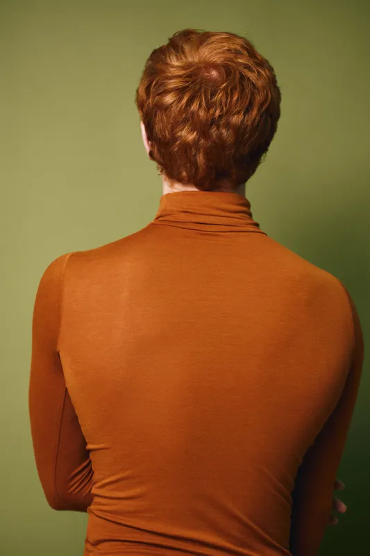 Redhead person with short hair wearing orange turtleneck sweater standing with back against olive background. Casual clothing, studio portrait, warm colors and minimal style.