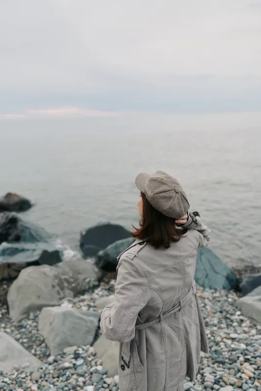 Woman in grey coat and checkered hat stands on rocky beach facing calm sea. Moody overcast sky creates peaceful, contemplative atmosphere. Concept of solitude and nature.