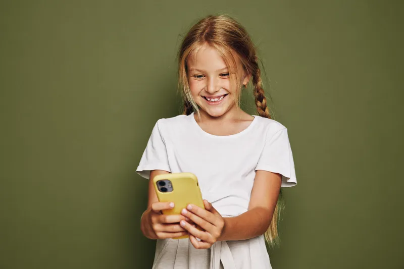 Smiling girl with braided hair wearing a white shirt looks at her yellow smartphone. She stands against an olive green background. Concept of childhood and technology.