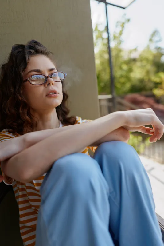 Young woman with curly hair sits against a wall in a striped top and blue pants, gazing thoughtfully into the distance on a sunny day with soft natural light and a calm mood
