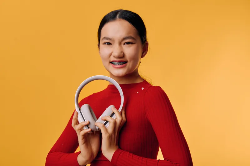 Young woman with braces smiling and holding gray headphones against yellow background, wearing red sweater with long sleeves and casual hairstyle for music enjoyment.