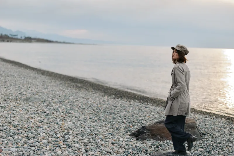 A woman in a grey coat and hat walks alone on a pebble beach near calm water under an overcast sky. The scene conveys solitude, contemplation, and peaceful nature.