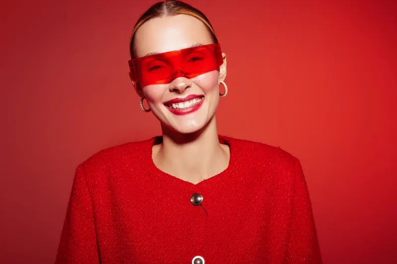 Smiling woman in red outfit and red translucent glasses poses against red background. Bright studio lighting highlights her joyful expression and modern fashion style. Concept of happiness.
