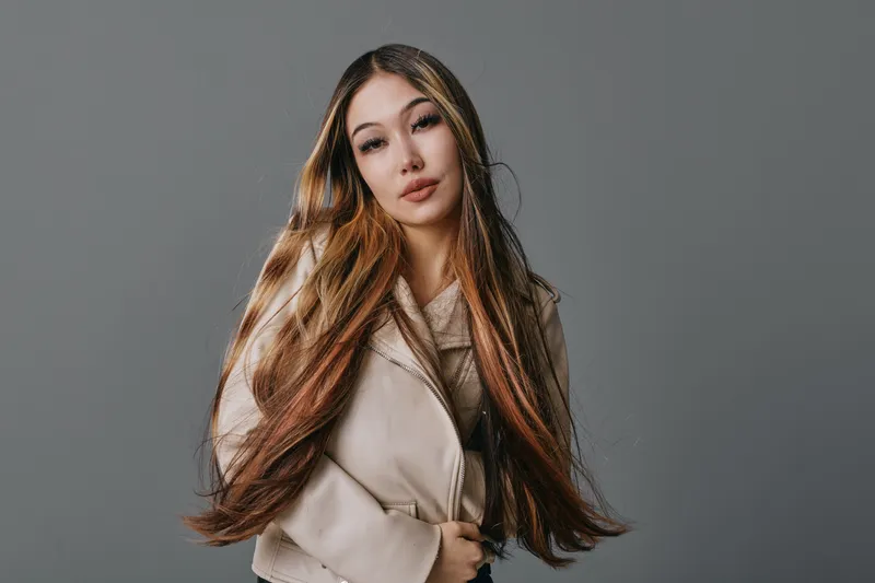 Young woman with long brown hair and neutral makeup wearing a beige coat posing with hands in pockets against gray background in studio portrait.