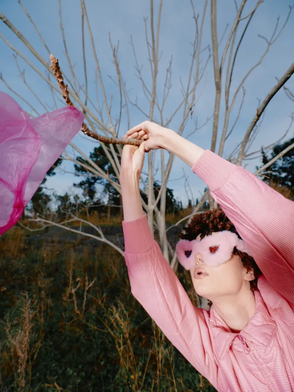 Woman in a pink sweater wearing a pink eye mask dances outdoors among bare trees, capturing a playful whimsical moment with flowing fabric and motion.