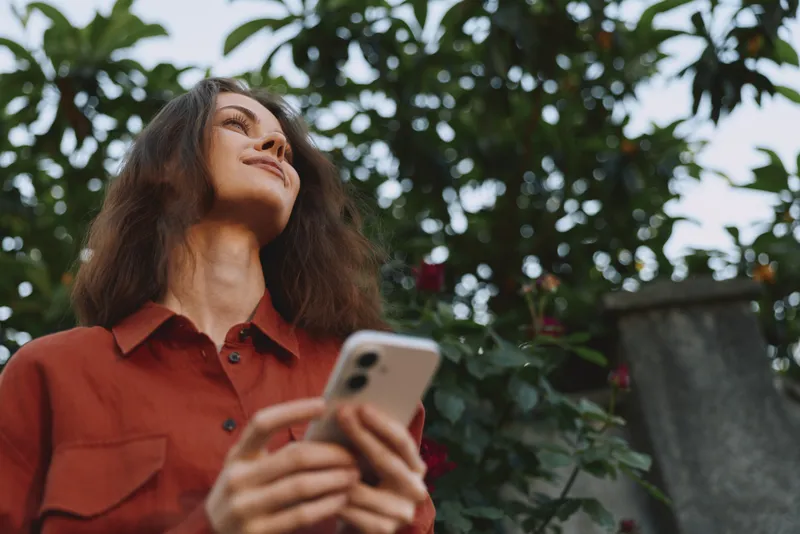 Smiling woman outdoors holding a smartphone, looking up with contentment and curiosity. Warm natural light highlights garden greenery and a relaxed, confident mood, suitable for lifestyle themes