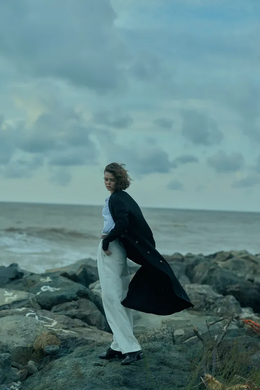 Wind swept coastal scene with a woman standing on rocks by the sea, wearing a long dark coat and light trousers, conveying motion and moody ocean atmosphere for dramatic mood