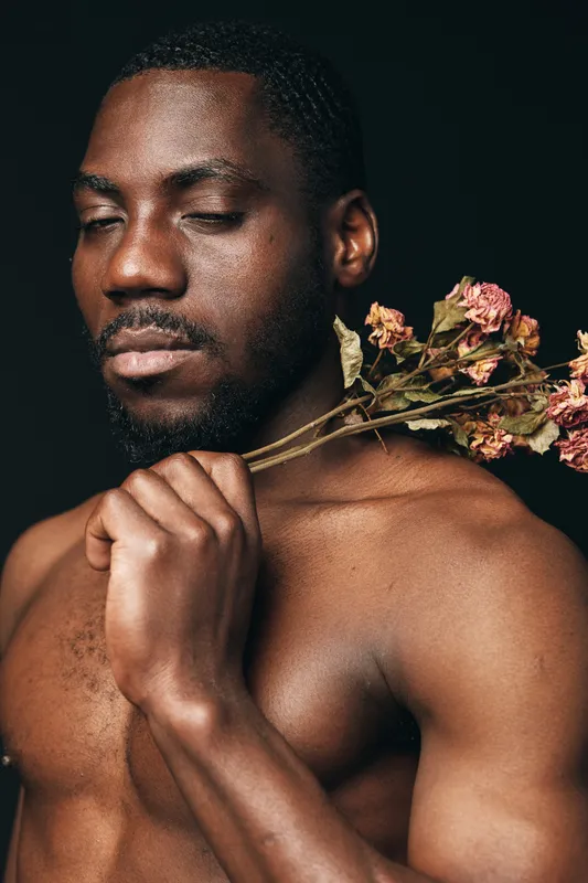 African man with closed eyes holding dried flowers on shoulder in creative conceptual visual style. Portrait of shirtless black male with thoughtful expression and artistic mood on dark background.