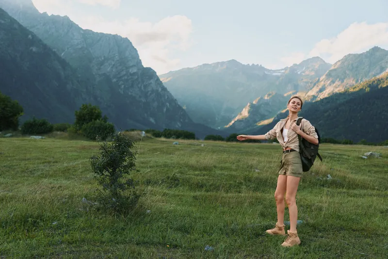 A young woman in hiking gear stands in a green valley between towering mountains, backpack slung over one shoulder, arms outstretched to feel the expansive scenery.
