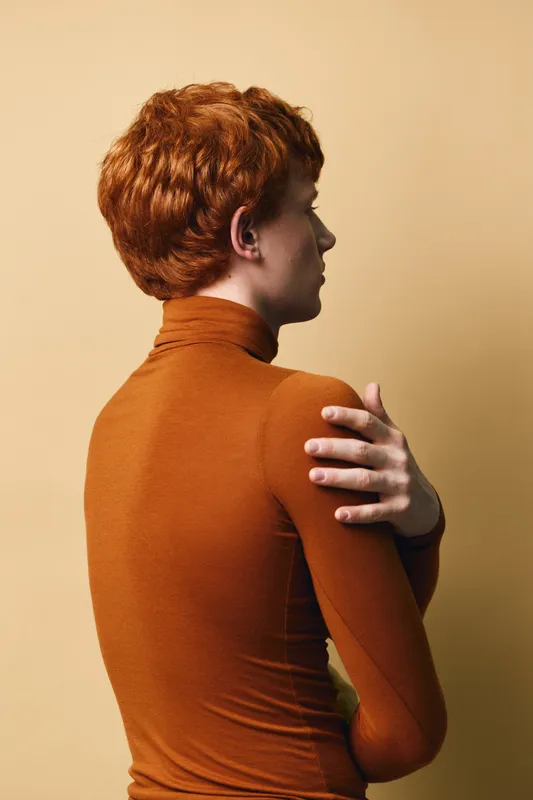 Portrait of redhead woman in brown turtleneck sweater standing sideways with crossed arms against beige background. Studio shot of thoughtful female with short hair and fair skin.