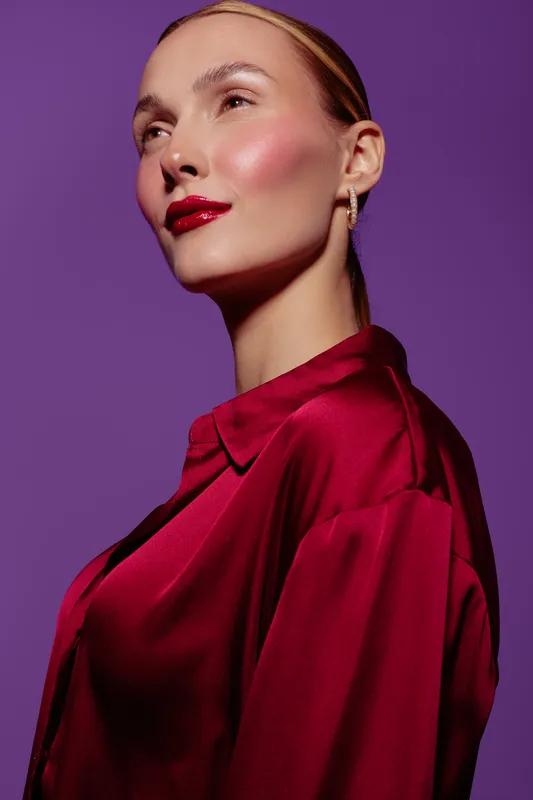 Portrait of confident woman with red lipstick and shiny satin blouse on purple background. Studio lighting highlights her smooth skin and elegant look. Concept of beauty and fashion.