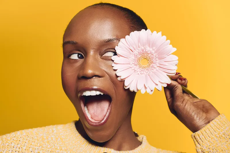 Happy African woman with dark skin holding a pink flower near her eye, smiling with open mouth on yellow background. Concept of joy, beauty, and natural skincare.