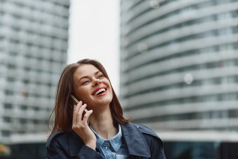Young woman with brown hair smiles happily while talking on a smartphone outdoors in an urban setting with modern buildings in the background.