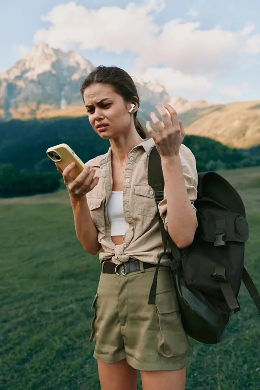 Young woman outdoors hiking with backpack looking confused at smartphone wearing casual clothes earbuds mountains background natural light