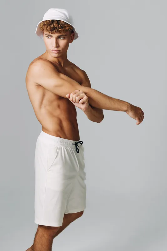 Young man with strong muscles wearing white shorts and bucket hat stretching his arm on gray background. Male fitness model preparing for training looking sideways with serious expression and cropped
