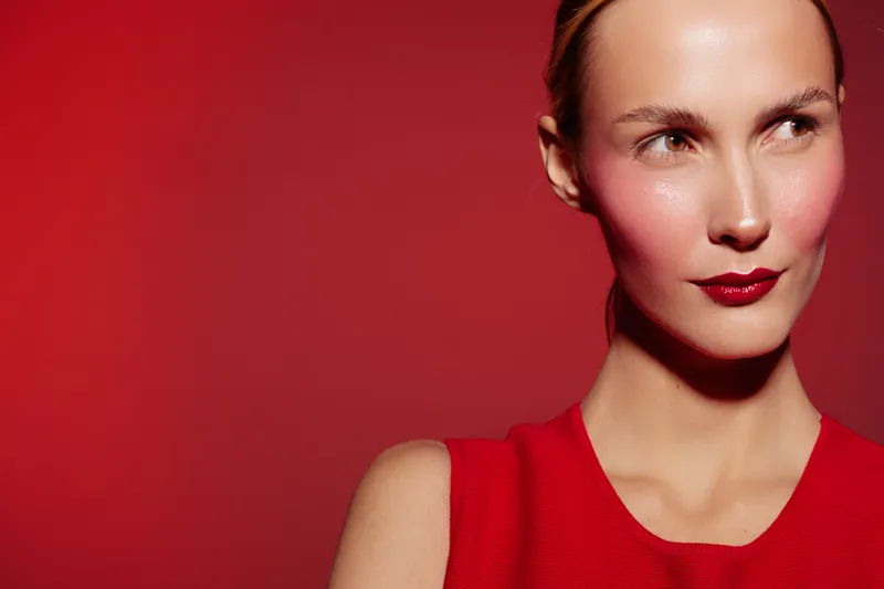 Close-up of a woman with red lipstick and blush wearing a red sleeveless top against a red background. Concept of beauty, makeup, fashion, and confident expression.