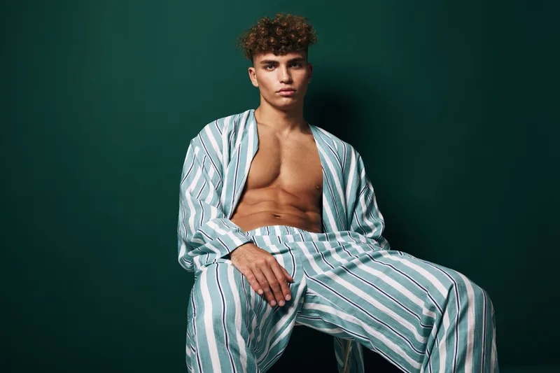 Young man with curly hair wearing striped shirt and pants sitting against green background looking confidently at camera with serious expression and casual style.
