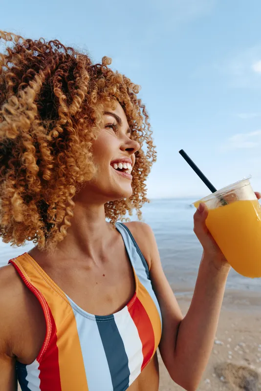 Woman with curly hair smiling on beach, ocean horizon behind, holding juice drink with straw, wearing swimsuit and colorful vest, outdoor summer vacation, sunny coastal wind