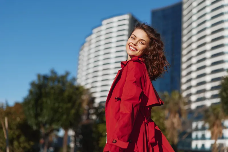 Young woman with curly hair wears a bright red trench coat and smiles confidently as she stands outdoors in an urban park with tall modern buildings behind, capturing a lively city moment.