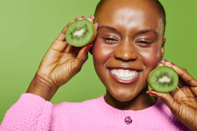 Happy smiling African woman with dark skin holds two halves of fresh kiwi fruit near face on green background. Concept for healthy lifestyle, natural food, wellness and beauty care.
