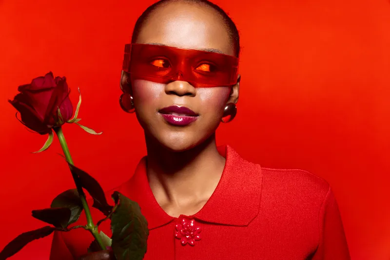 Portrait of a dark skinned African woman wearing red sunglasses and red outfit holding a red rose. Studio shot with red background. Concept of fashion, beauty, and elegance.