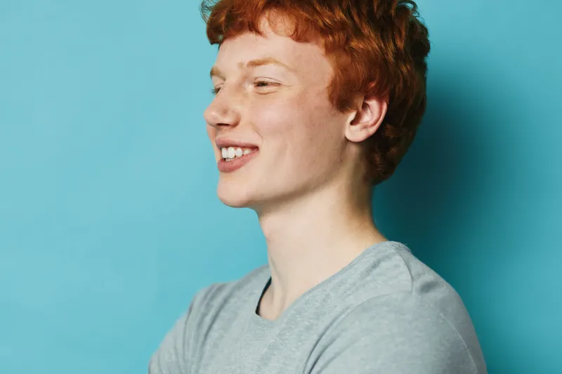 Smiling young man with red hair wearing gray shirt posing sideways against blue background with closed eyes and cheerful expression in casual studio portrait.