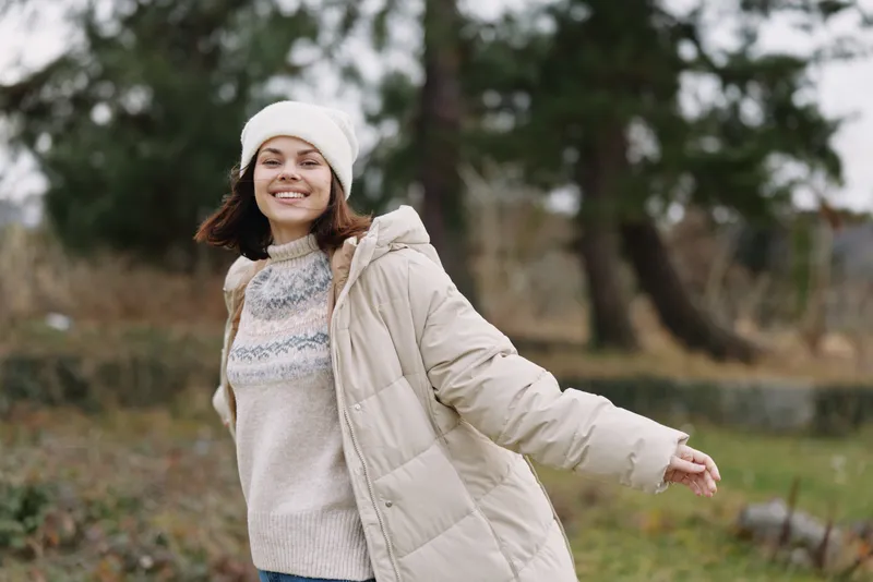 Young woman in a warm coat and beanie outdoors, smiling as she enjoys a crisp autumn day in a park with trees, leaves, and soft sunlight