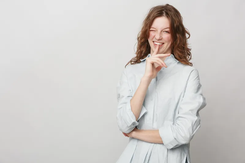 Smiling young woman with curly hair wearing white shirt holds finger near lips in shushing gesture. Studio portrait on light background. Concept of secrecy, silence, and playful mood.