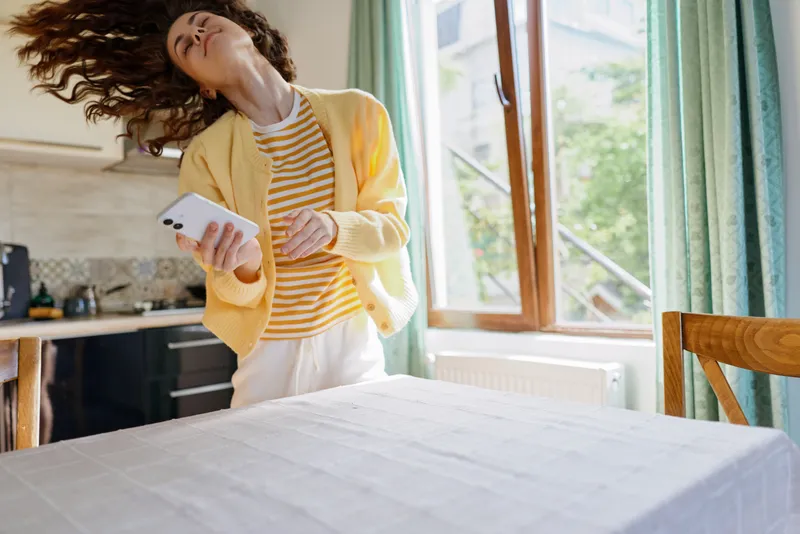 A woman in a yellow cardigan and striped top dances by a sunlit window, phone in hand. A bright home interior creates a joyful, relaxed mood for everyday living.