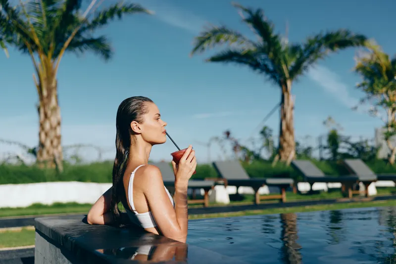 Relaxed woman in a white swimsuit lounges at a rooftop pool under clear sky, sipping a drink with straw, enjoying warm sunshine and tropical ambience