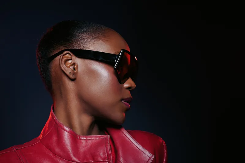 Profile of a confident black woman with dark skin wearing large sunglasses and a red leather jacket against a dark background. Concept of fashion, style, and attitude.
