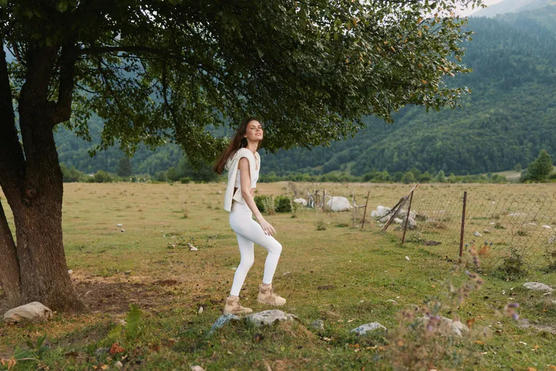 Woman in white outfit walking through a grassy field beside a large tree, mountains in the distance. Calm outdoor lifestyle shot emphasizing nature and exploration.