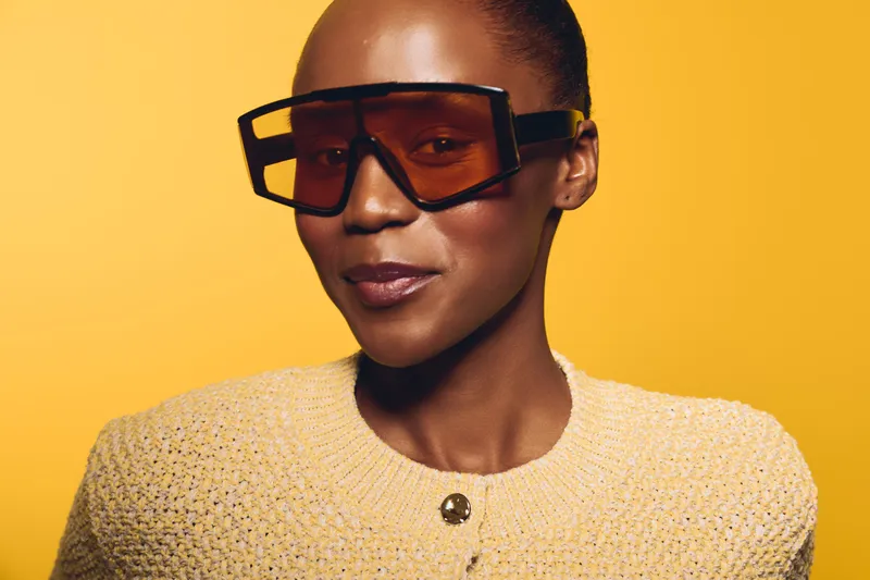 Confident African woman with dark skin wearing oversized sunglasses and a knitted sweater poses against a yellow background. Concept for fashion, style, and beauty portrait.