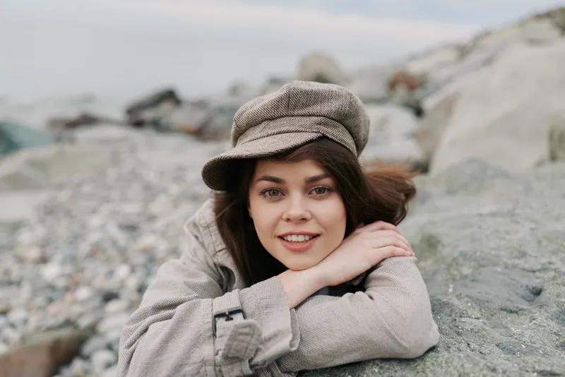 Young woman with brown hair wearing checkered cap and coat lies on rocky beach smiling. Natural outdoor portrait with casual autumn fashion and relaxed mood by the sea.