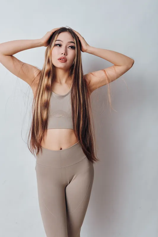 Young woman with long hair wearing beige sportswear poses with hands on head against plain wall. Fitness outfit, casual style and natural makeup for healthy lifestyle and beauty.