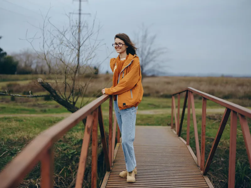 Woman lifestyle outdoor walking on a wooden bridge in countryside, orange jacket and jeans, calm face with glasses, cloudy sky and spring fields, peaceful mood solo travel inspiration
