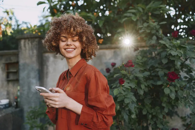 Smiling young woman with curly hair in a rust red blouse checks her phone in a sunlit garden, capturing a candid lifestyle moment of connection and calm