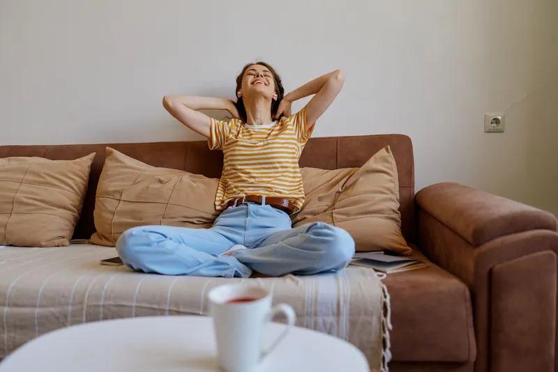 Relaxed woman on a plush sofa lounges with arms behind her head, wearing a striped shirt and jeans, soft lighting creates a warm cozy living area with cushions and daylight indoors today