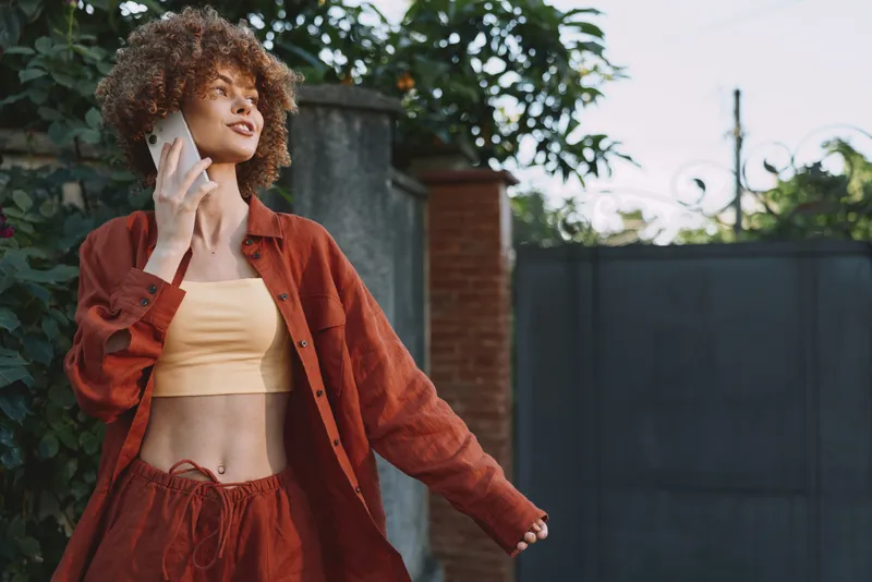 Smiling woman with curly hair wearing an orange jacket and yellow top outdoors. Casual lifestyle, fashion, confidence, and sunshine create a warm, inviting mood