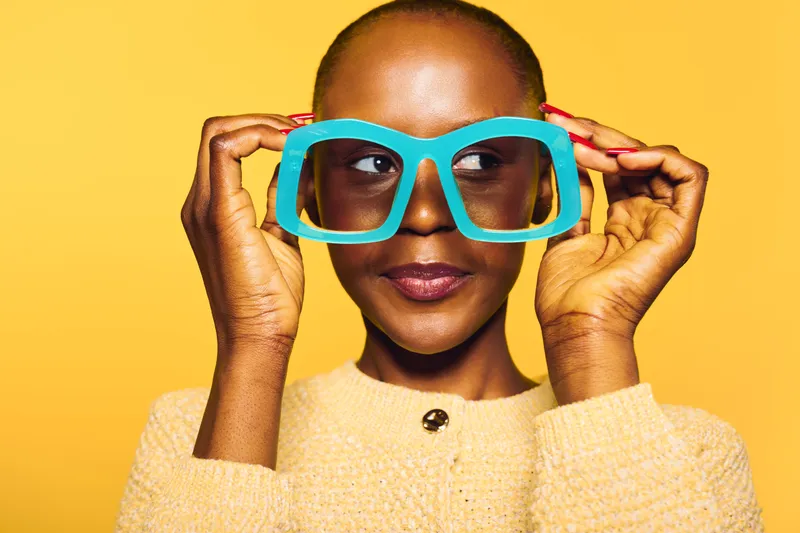 Happy African woman with dark skin holds large blue glasses near face, wears yellow sweater on yellow background. Concept of fashion, fun, and vibrant summer style.