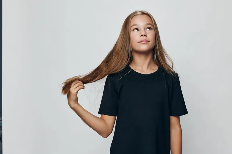 Girl with long brown hair wearing black t shirt holds hair and looks up on gray background. Portrait of child with thoughtful expression and casual style in studio.