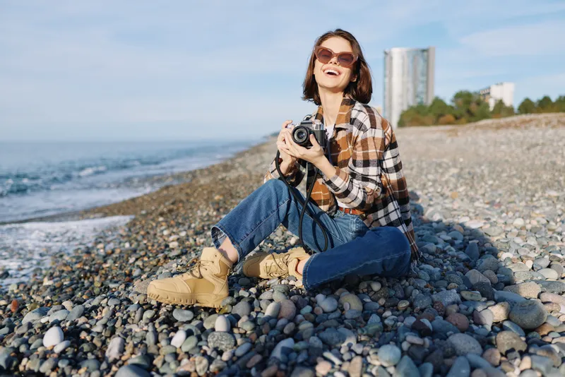 Casual woman photographer sits on a pebbled beach holding a camera, wearing plaid shirt and jeans, enjoying a sunny seaside moment with distant buildings and calm blue sea.