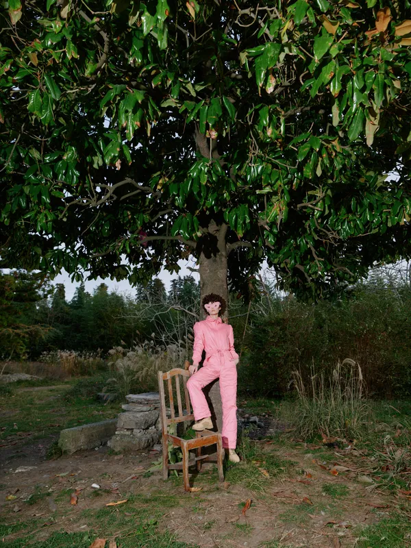 Child in a pink outfit stands on a wooden chair under a large leafy tree in an outdoor garden, creating a playful portrait with natural light and rustic surroundings
