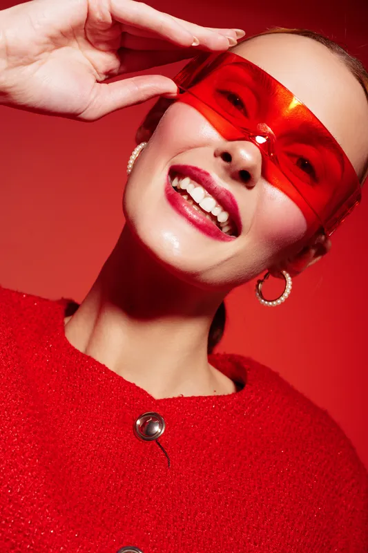 Smiling woman in red sweater and red translucent glasses poses against red background. Close-up portrait with bright makeup and hoop earrings. Concept of fashion, style, and vibrant mood.