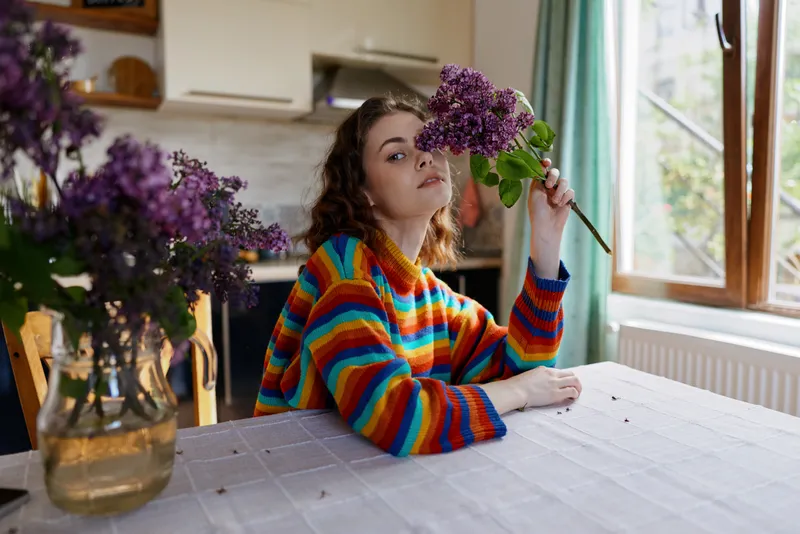Cheerful woman indoors, wearing a colorful striped sweater, holding a purple bouquet by a sunny kitchen window, home lifestyle moment, soft natural light, cozy atmosphere with warm tone