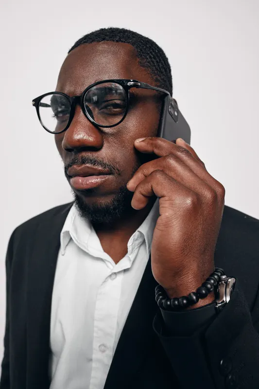 African man with dark skin wearing glasses and black suit talking on smartphone in studio. Closeup portrait of confident businessman with beard and white shirt on white background.