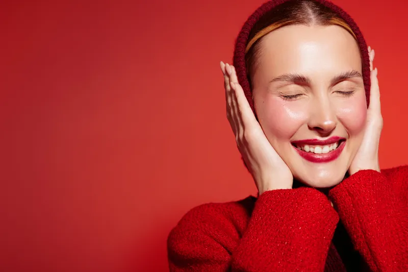 Smiling woman with closed eyes wearing red sweater and headband touches face with hands. Bright red background creates warm and joyful mood. Concept of happiness, beauty, and self-care.