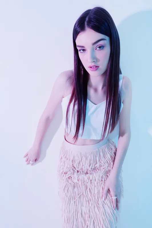 Young woman with long dark hair wearing a white top and fringed skirt posing in soft pastel lighting. Concept of woman beauty fashion with a modern stylish look.