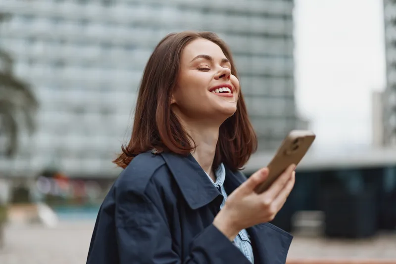 Young woman with brown hair smiles joyfully while holding a smartphone outdoors in an urban environment wearing a dark coat and light shirt.
